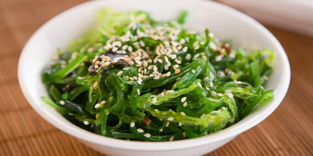 Sałatka z glonów wakame, posypana sezamem, w białej miseczce na bambusowej macie. Idealna na letnie orzeźwienie.