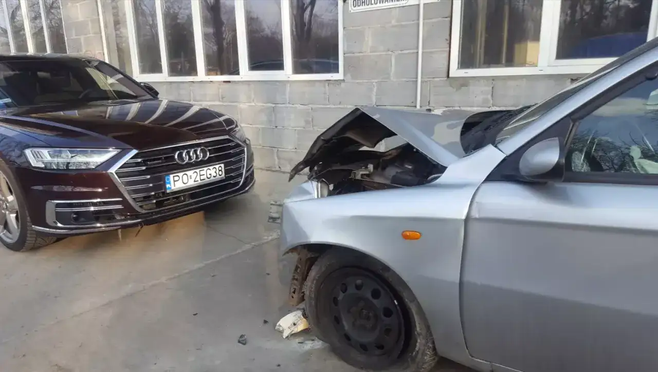 Uszkodzony srebrny samochód, którego maska jest otwarta, obok stoi ciemny Audi. Szkoda z OC sprawcy, auto w leasingu.