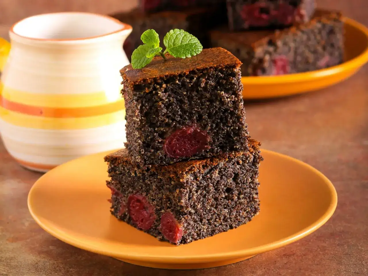 Wilgotny makowiec z gotowej masy makowej, z wiśniami, udekorowany miętą, na pomarańczowym talerzu.