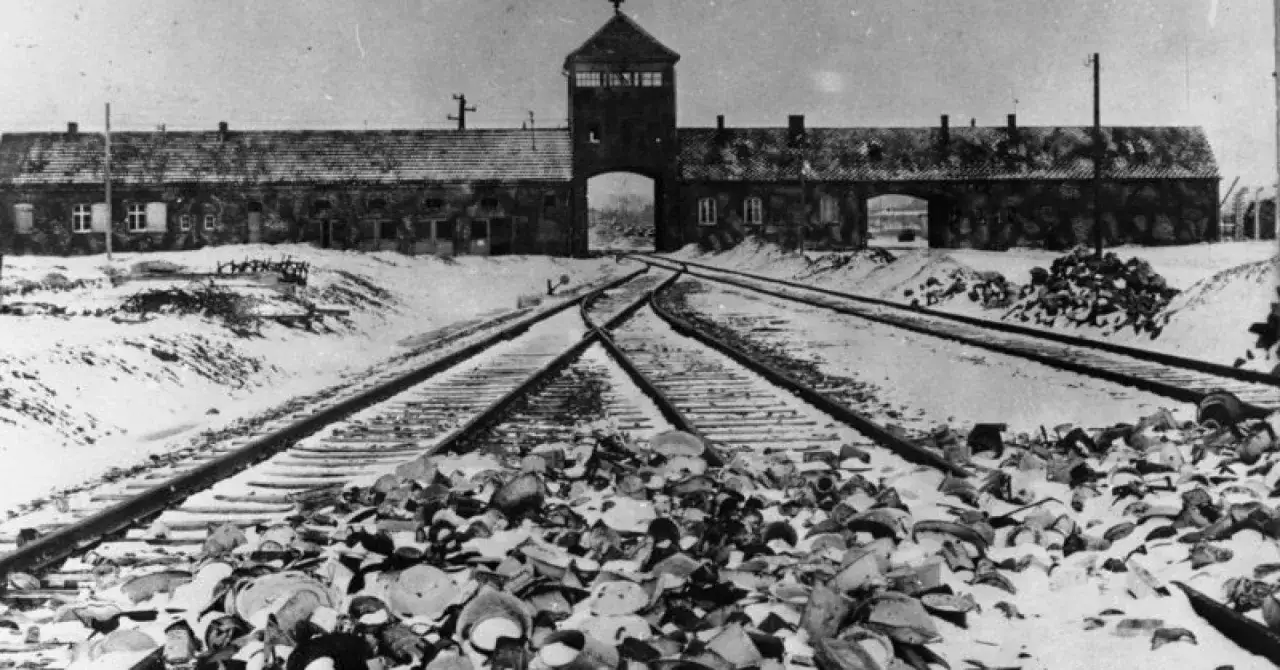 Brama obozu Auschwitz w zimowej scenerii, z torami kolejowymi i stertą przedmiot&oacute;w. Obraz przypomina o tragedii, o kt&oacute;rej czytamy w książkach o Auschwitz.