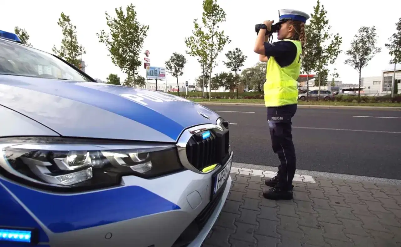Policjantka w kamizelce odblaskowej obserwuje ruch przez lornetkę, stojąc obok radiowozu. Czeka na okazję do jazdy na zderzaku.
