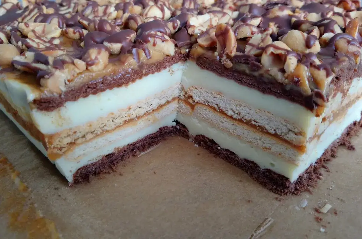 Pyszne ciasto snickers bez pieczenia z warstwami kremu, ciastek i karmelu, posypane orzeszkami i polane czekoladą.