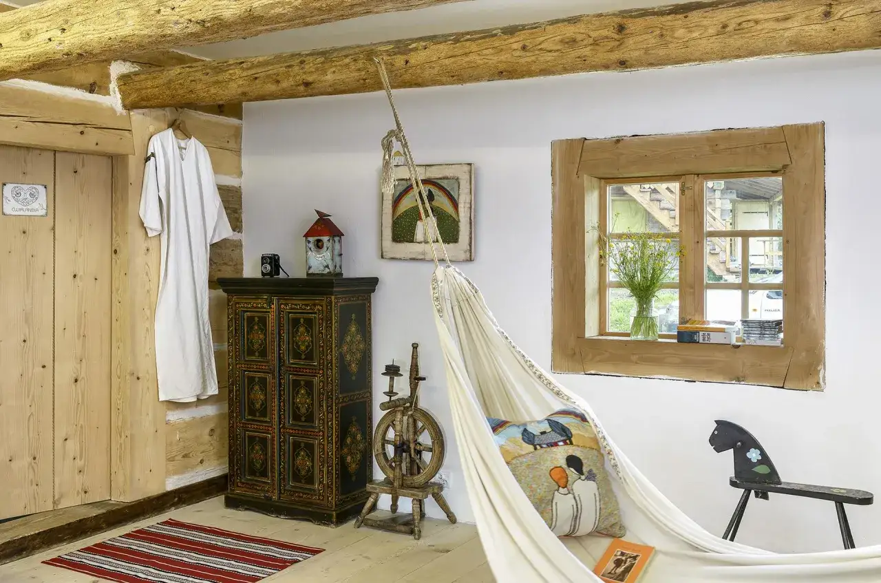 Przytulne wnętrze domu w stylu wiejskim: biała hamak, zdobiona szafa, kołowrotek i drewniane belki tworzą sielski klimat.