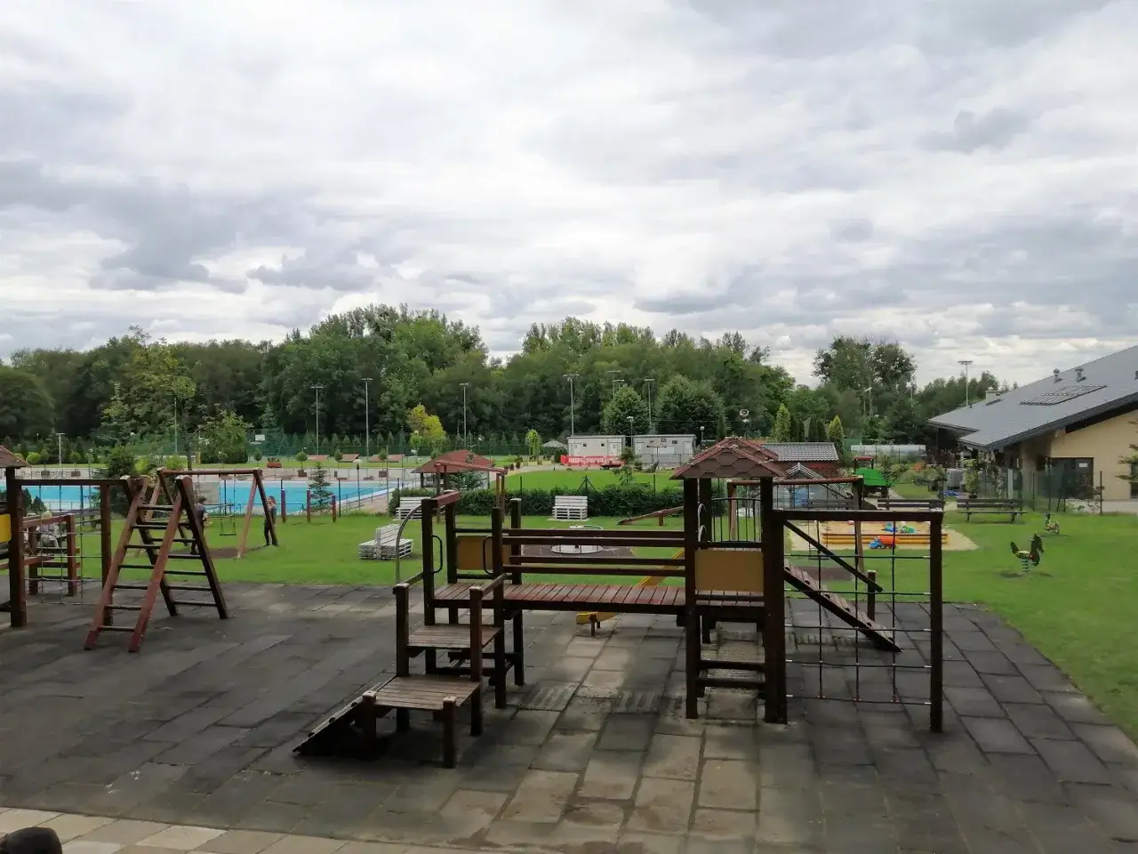 Plac zabaw z basenem w tle, w pobliżu lasu. Widać zjeżdżalnie, huśtawki i piaskownicę. Miejsce idealne na rodzinny wypoczynek, jak na Księżej Górze w Radzionkowie.