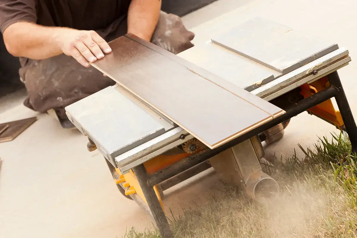 Ręce mężczyzny precyzyjnie prowadzą panel winylowy do cięcia na pile stołowej. Kurz wzbija się w powietrze, tworząc atmosferę pracy.