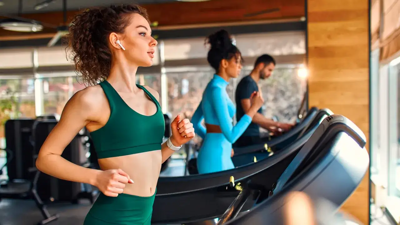 Kobieta w zielonym sportowym topie biegnie na bieżni, słuchając muzyki. Dowiedz się, jak włączyć bieżnię na siłowni, by zacząć trening.
