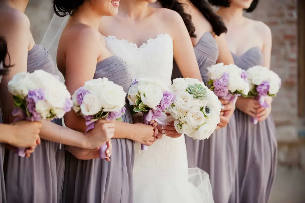Uśmiechnięte druhny w szarych sukniach trzymają bukiety dla świadkowej. Białe r&oacute;że i fioletowe kwiaty tworzą piękną kompozycję.