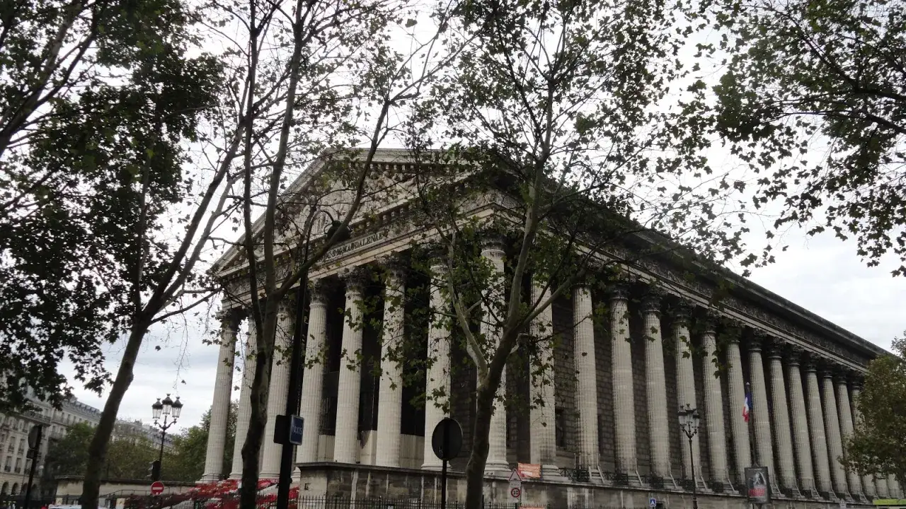 Majestatyczny kościół de la Madeleine w Paryżu, otoczony drzewami, z kolumnami w stylu korynckim.