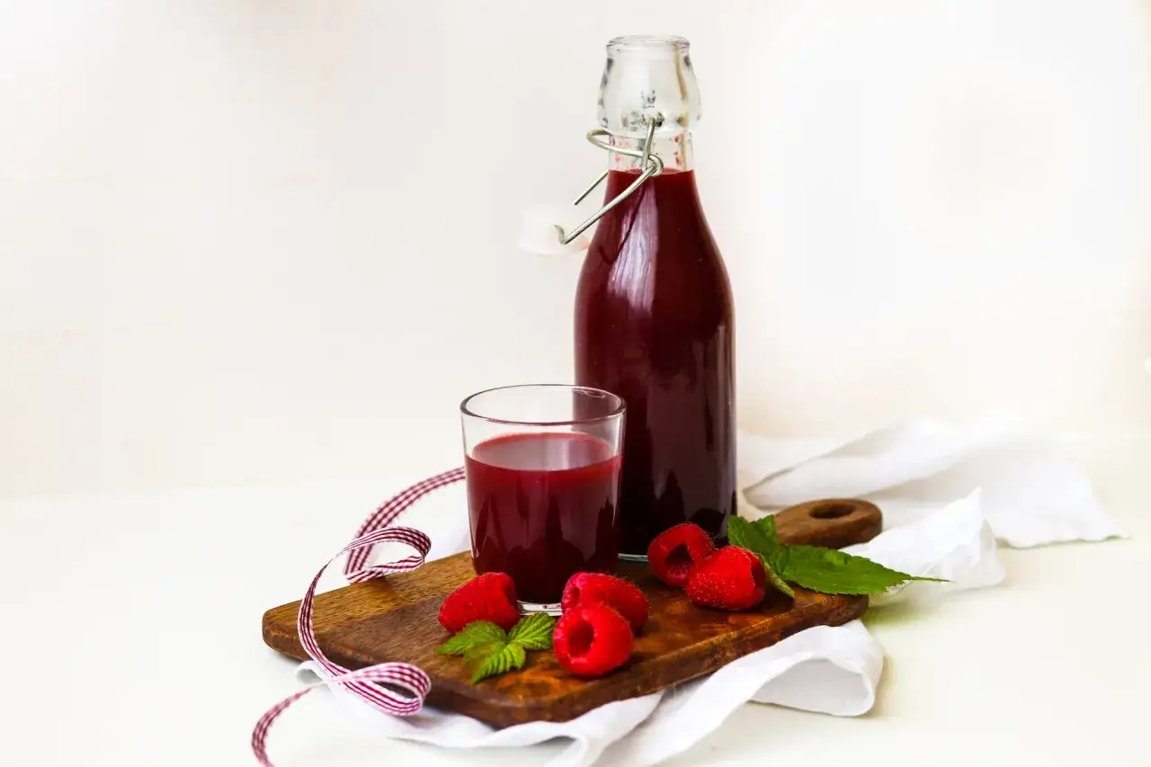 Sok z malin przepis babci: butelka i szklanka pełne pysznego, domowego soku, świeże maliny i listki mięty na desce.