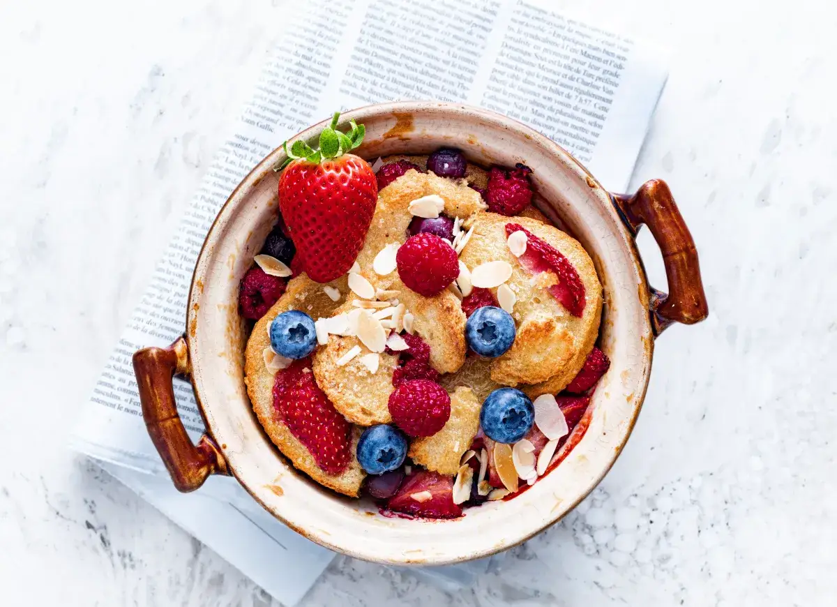 Pudding chlebowy z jagodami i truskawkami, posypany płatkami migdałów. Obok gazeta.