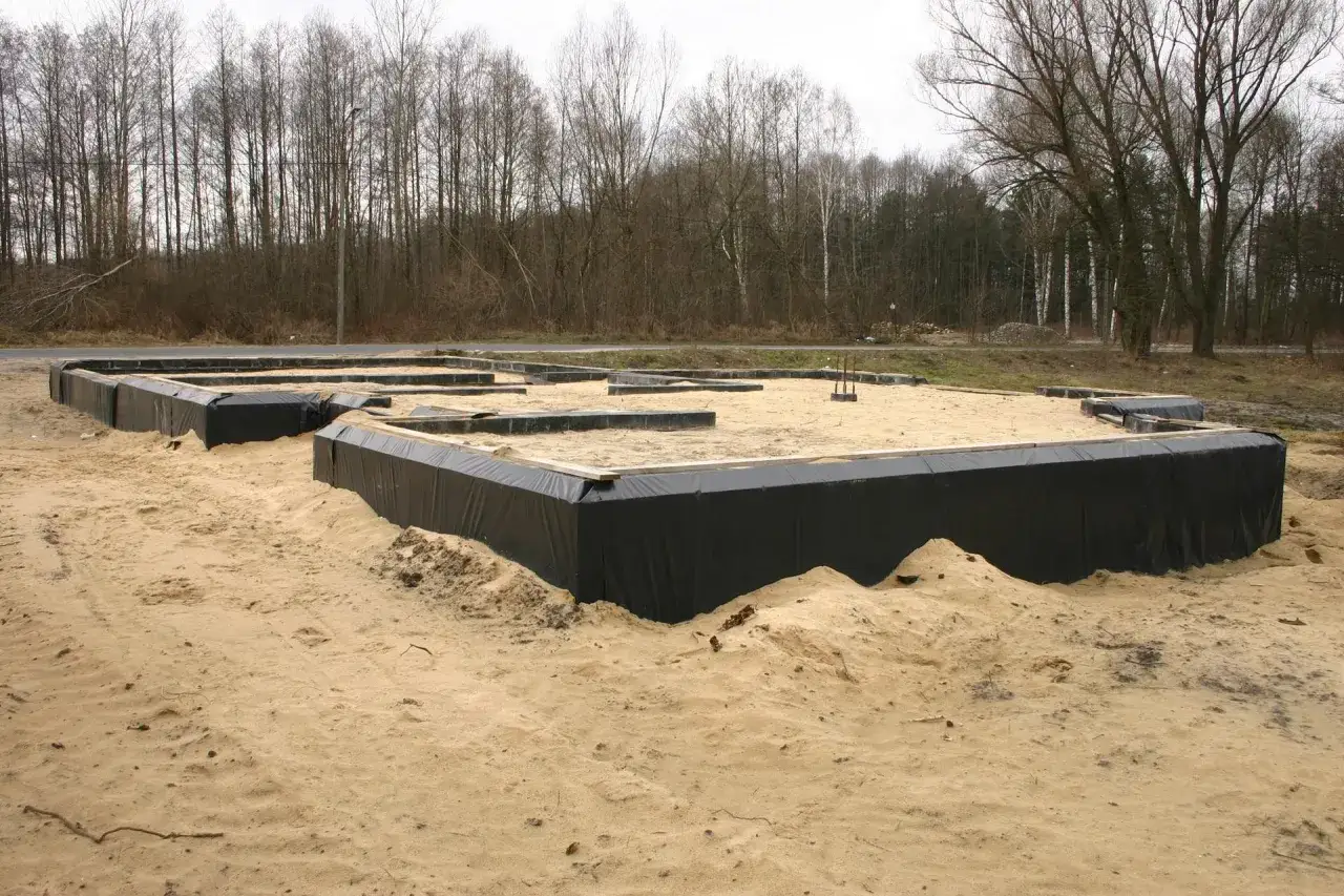 Fundament domu w piasku, otoczony czarną folią. To początek budowy, gdzie widać przygotowanie pod przyszły drenaż.