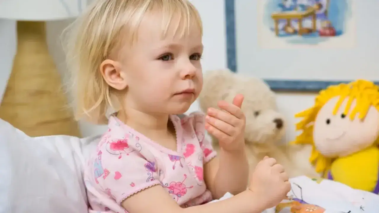 Mała dziewczynka z blond włosami, w r&oacute;żowej piżamce, siedzi w ł&oacute;żku z pluszakami. Martwi się długotrwałym kaszlem u dziecka bez zmian osłuchowych.