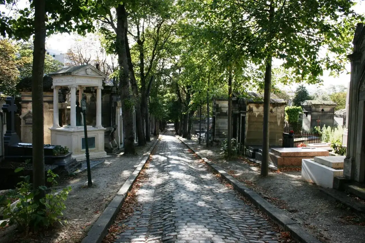 Aleja na cmentarzu Père Lachaise, wybrukowana kamieniem, otoczona drzewami i nagrobkami.