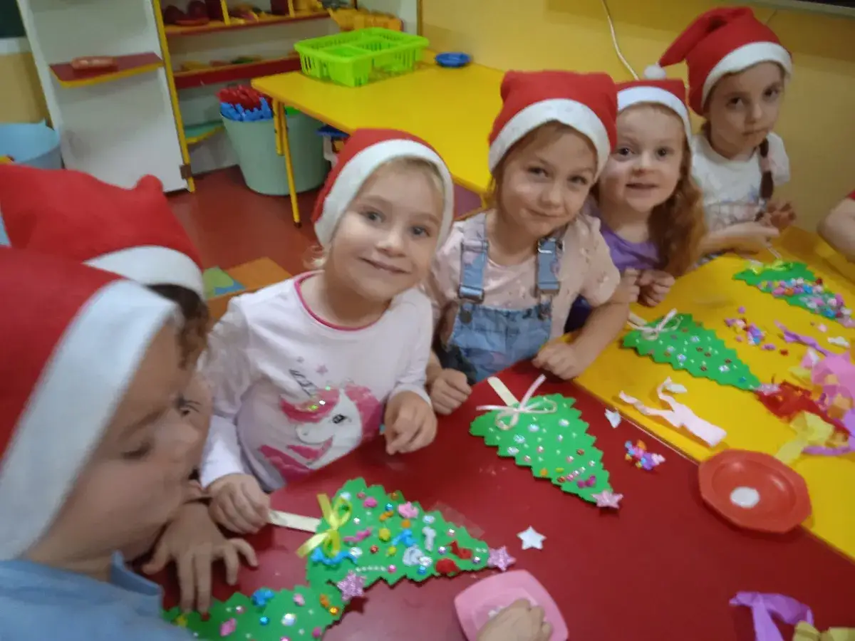 Wesołe **święta w przedszkolu**! Dzieci w mikołajkowych czapkach tworzą własne choinki z papieru i ozd&oacute;b.