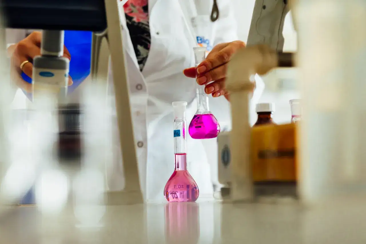 W laboratorium mikrobiologicznym pracownik w białym fartuchu trzyma kolbę z r&oacute;żowym płynem.