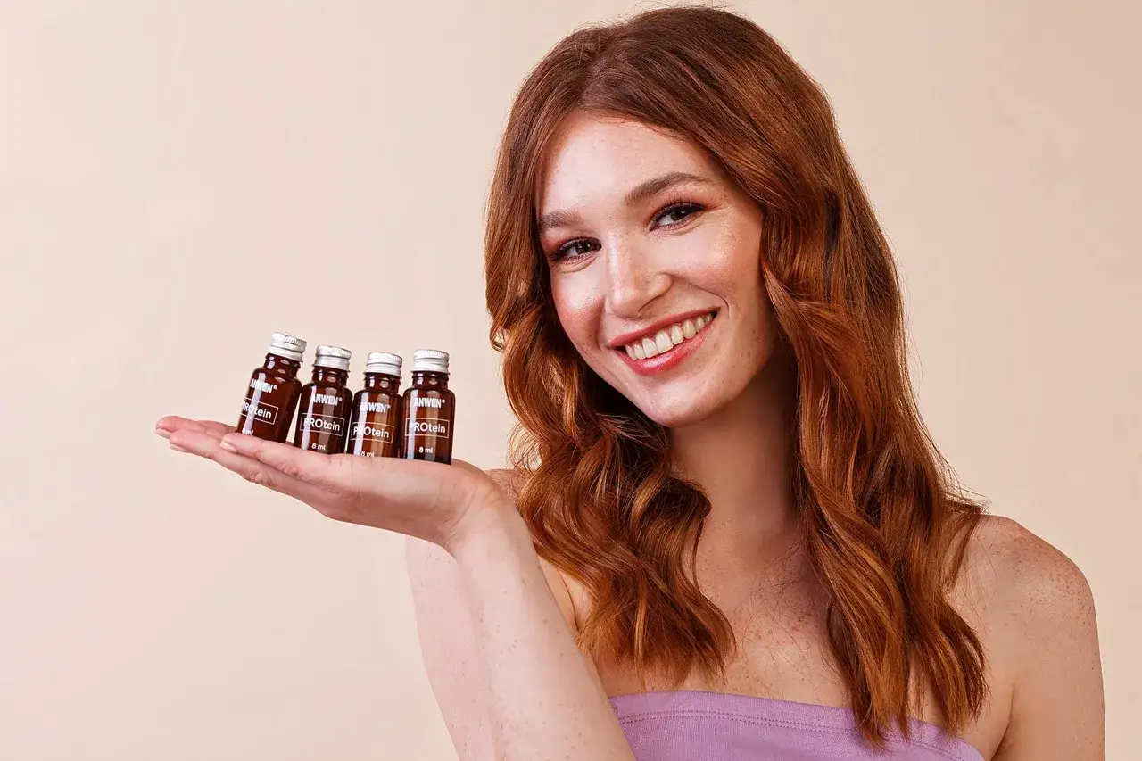 Uśmiechnięta rudowłosa kobieta prezentuje na dłoni cztery małe buteleczki z odżywczym serum. Dowiedz się, jak nakładać serum na włosy dla najlepszych efektów.