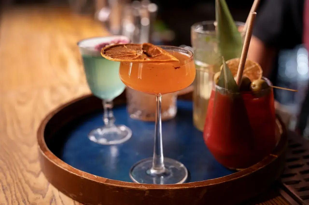 Pomarańczowy drink z Metaxą, ozdobiony suszoną pomarańczą, na tacy z innymi koktajlami.