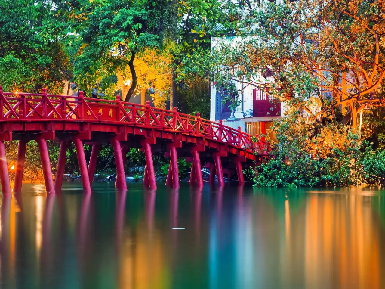 Czerwony most nad wodą, otoczony zielenią i drzewami w ciepłych barwach. Marzysz o takiej wycieczce do Wietnamu?