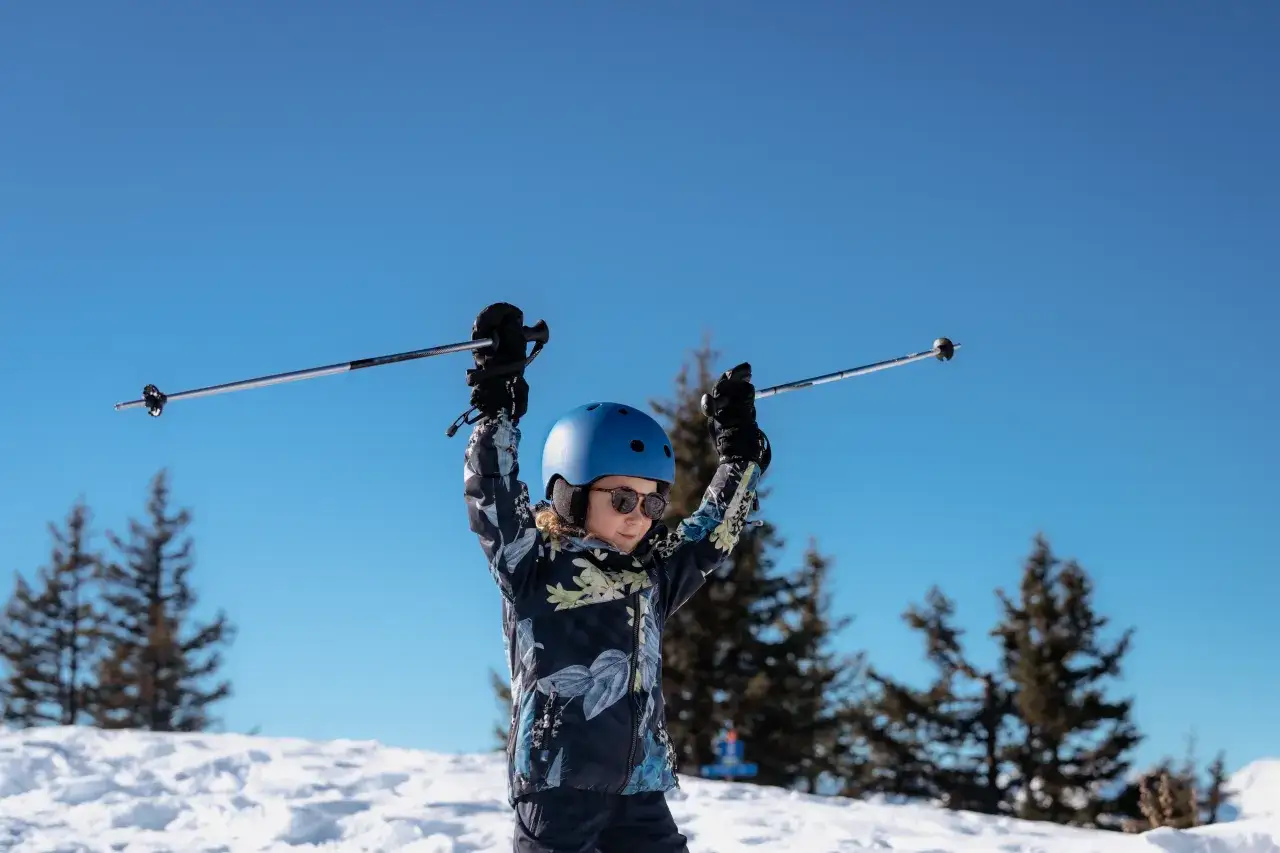 Mały narciarz w niebieskim kasku i kurtce z motywem kwiatowym, z radością unosi ręce z kijkami narciarskimi. Idealne narty dla 3 latka na pierwsze zjazdy.