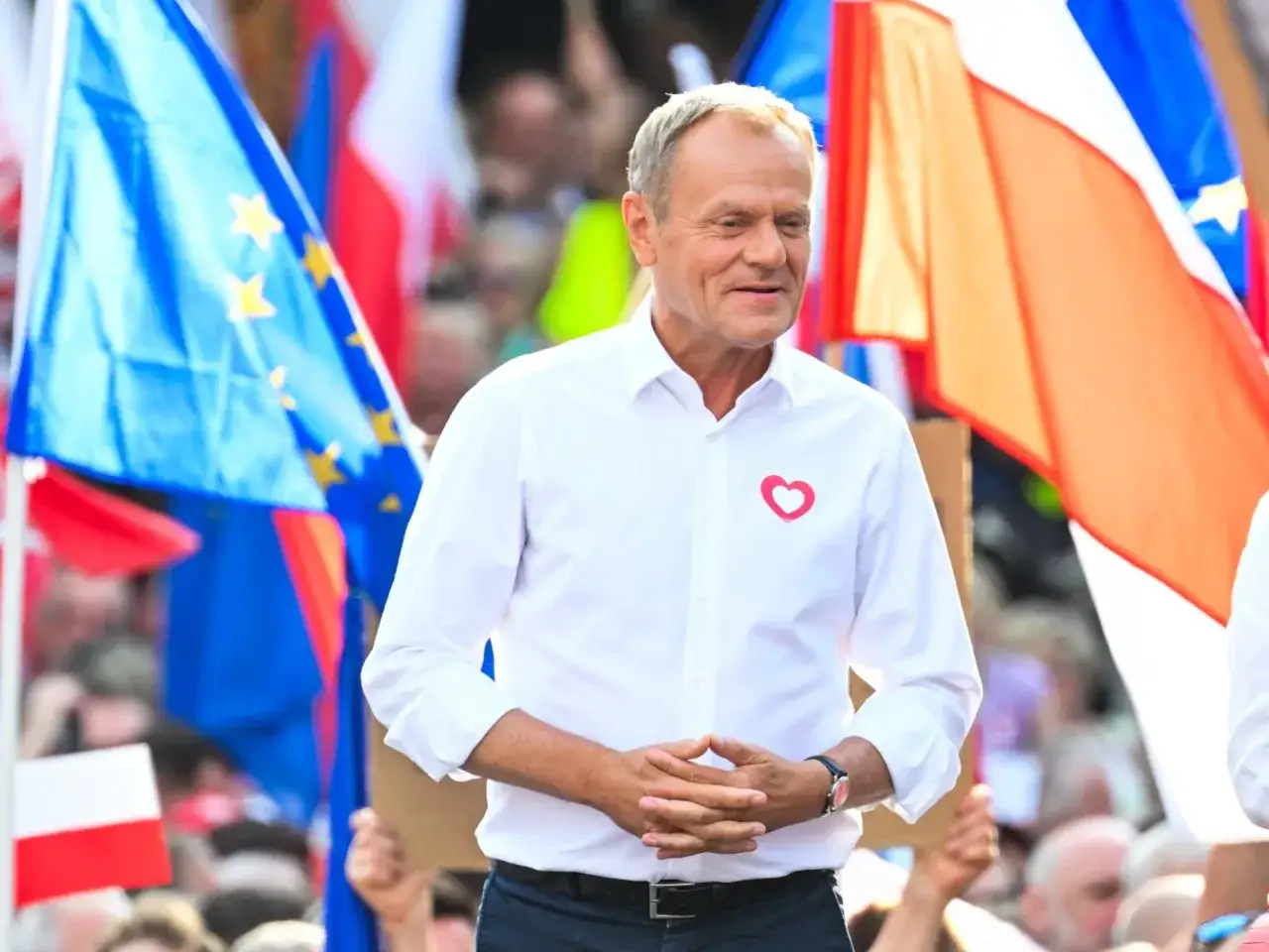 Donald Tusk w Ostródzie, na tle flag Polski i UE, z sercem na koszuli.