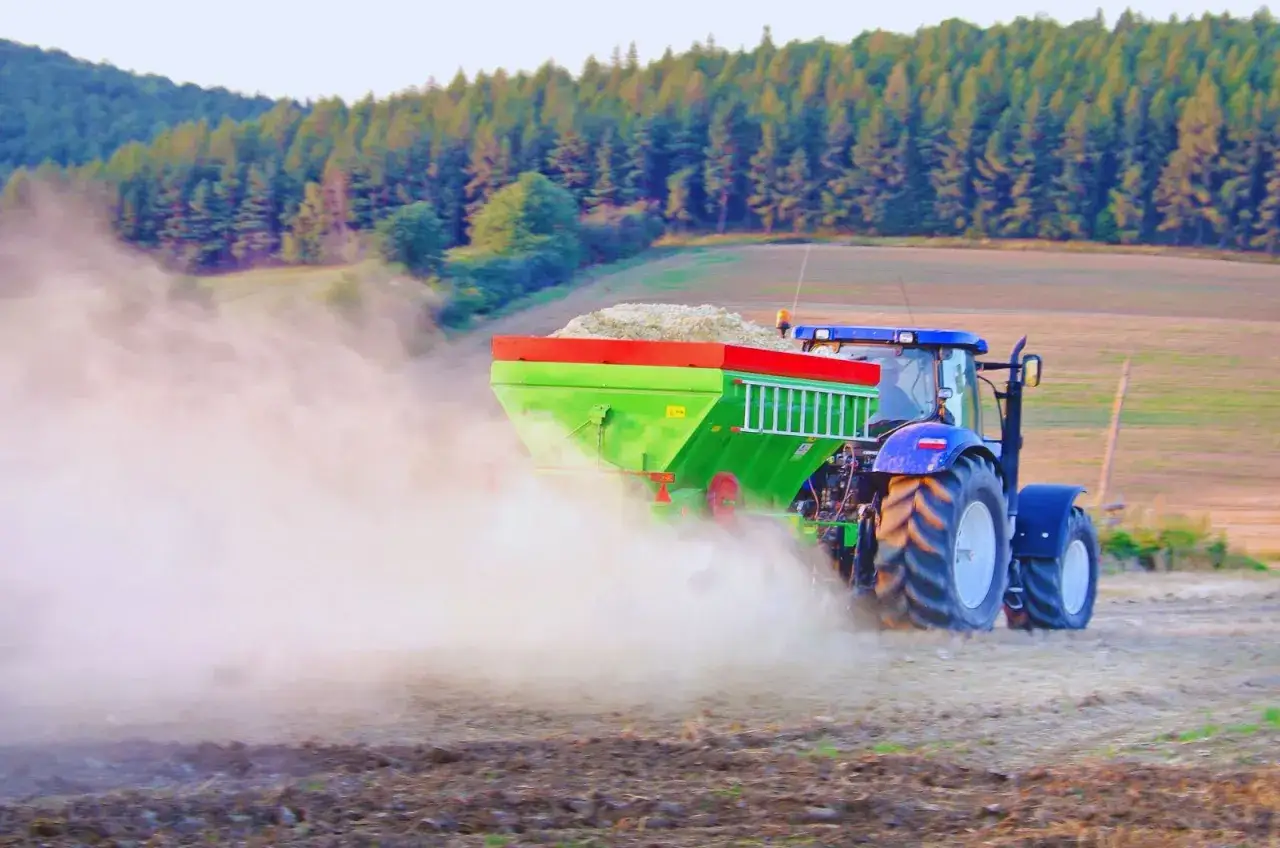 Traktor rozsiewa wapno w płynie na polu, tworząc chmurę pyłu.