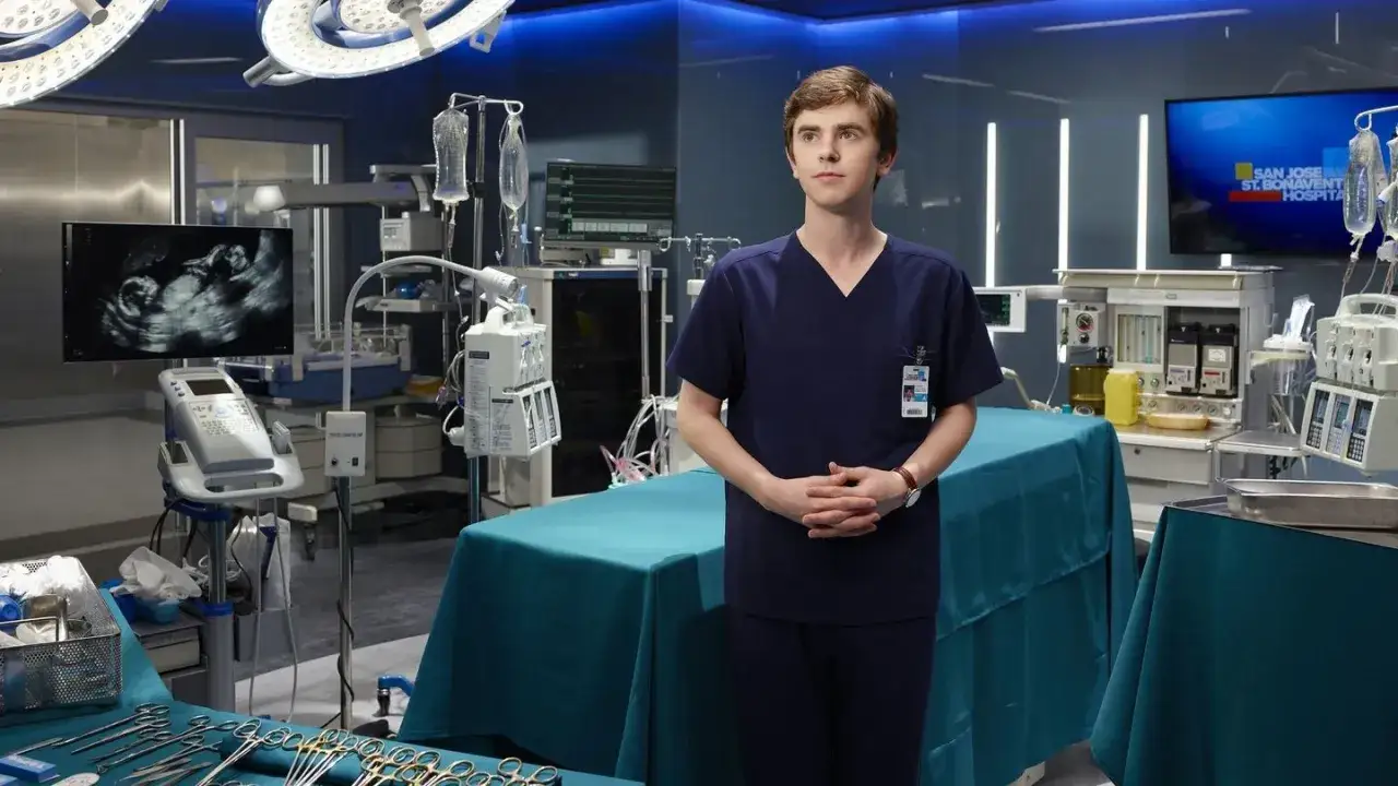 Młody lekarz w granatowym uniformie stoi na sali operacyjnej. W tle USG płodu i sprzęt medyczny.