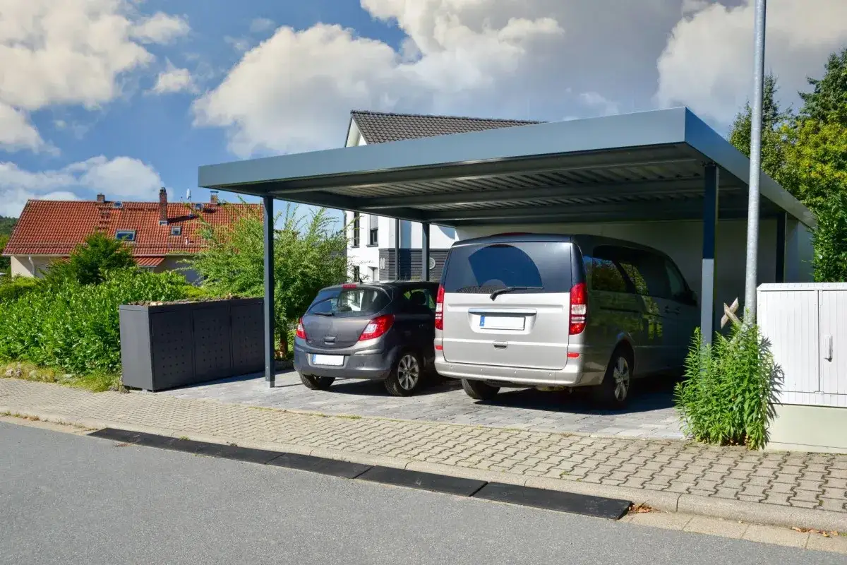 Nowoczesna wiata w granicy działki chroni srebrnego vana i szare auto. W tle domy i zieleń.