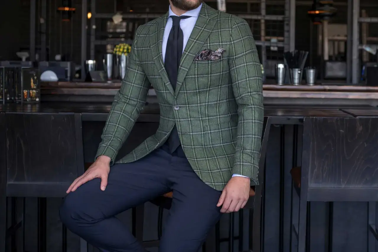 Mężczyzna w marynarce w kratę w kolorze butelkowa zieleń, z granatowymi spodniami i czarnym krawatem, siedzi przy barze.