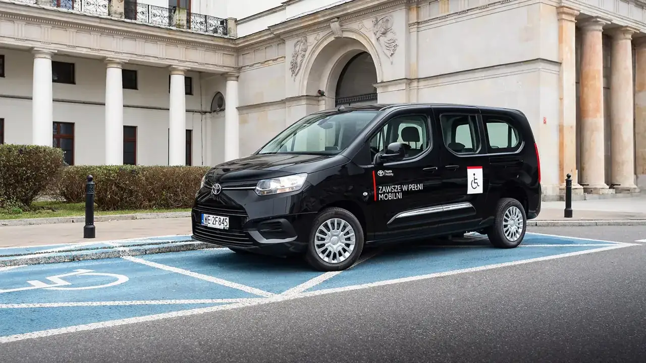 Czarny van Toyota z oznaczeniem dla niepełnosprawnych na niebieskim miejscu parkingowym. Dowiedz się, jak dostać dofinansowanie na samochód.