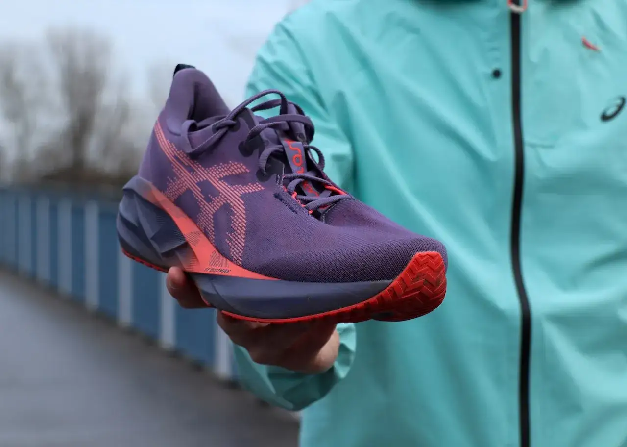 Fioletowe buty do biegania Asics Megablast z pomarańczowymi akcentami, trzymane w dłoni na tle mostu.