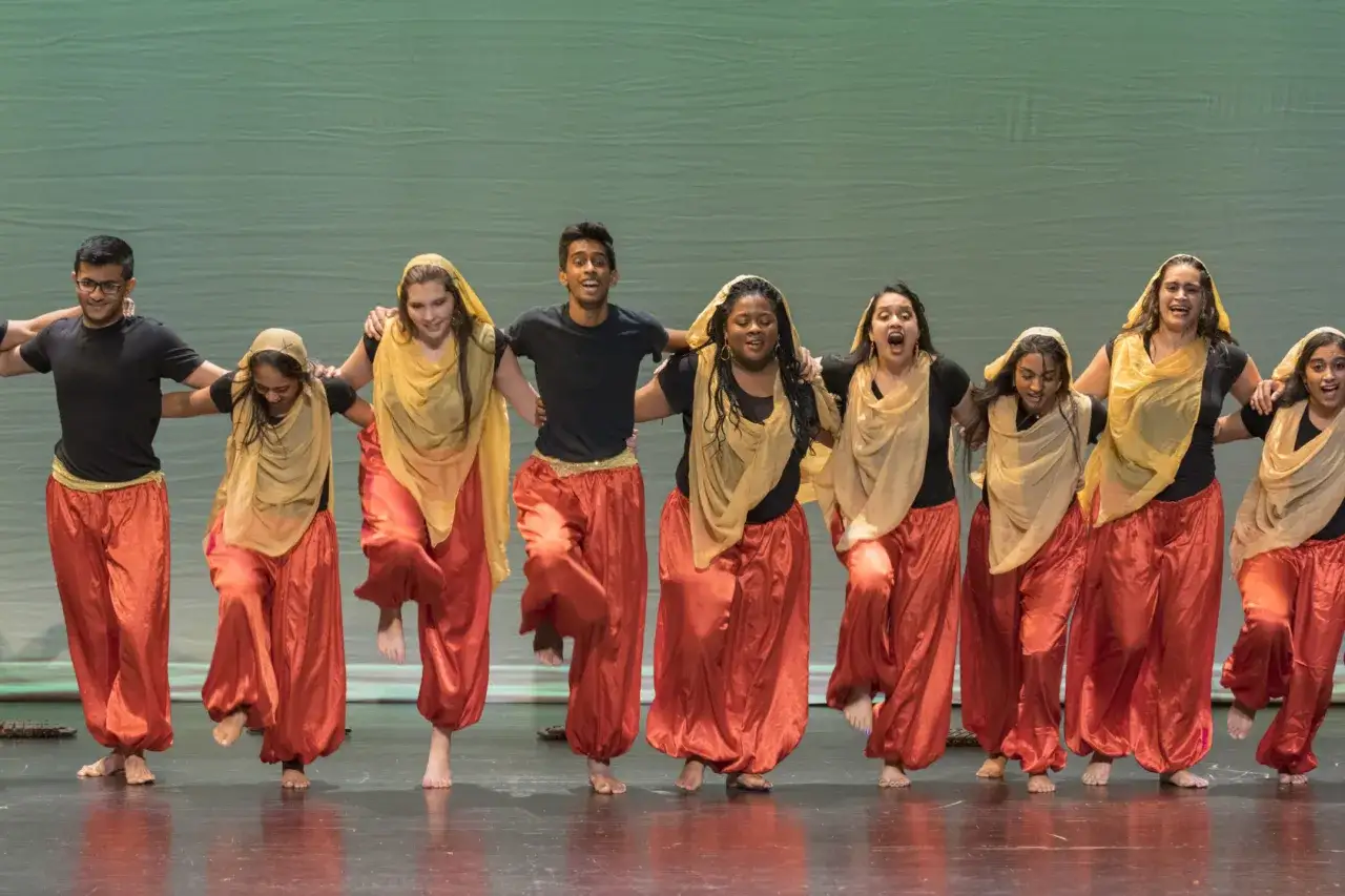 Grupa tancerzy w pomarańczowych spodniach i beżowych chustach wykonuje jeden z najpopularniejszych tańc&oacute;w świata.