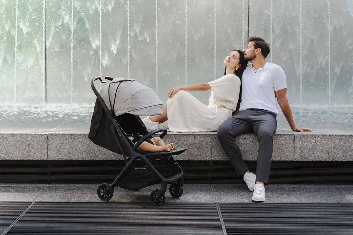 Para, siedząc przy fontannie, z dzieckiem w w&oacute;zku spacerowym. Czas na spacer&oacute;wkę, gdy dziecko zaczyna siedzieć.