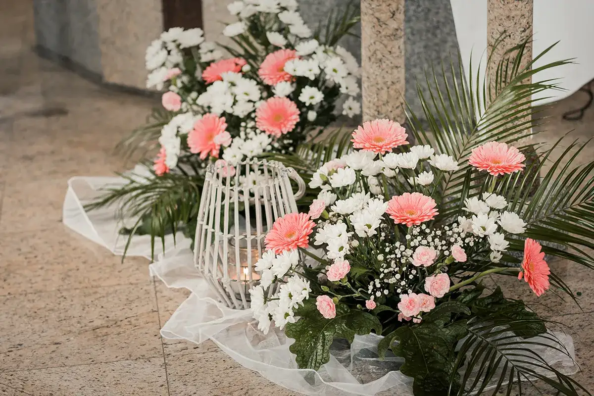 Wystrój kościoła na ślub: bujne bukiety różowych gerber i białych chryzantem, ozdobione zielenią, z lampionem.