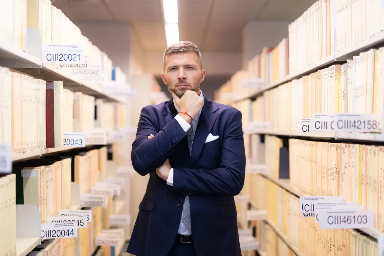 Mężczyzna w garniturze zastanawia się, kiedy złożyć wniosek o adwokata z urzędu, stojąc wśród regałów z dokumentami.