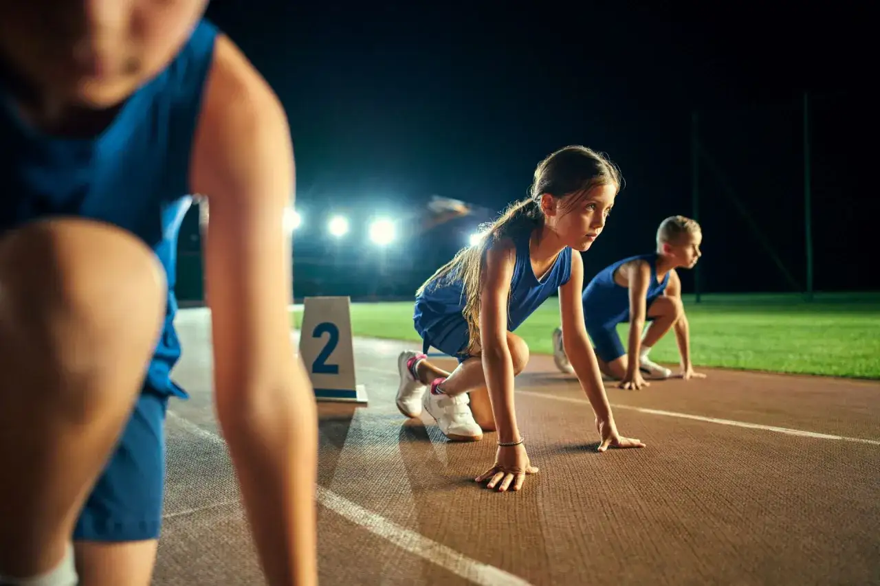 Dzieci w strojach sportowych na bieżni, gotowe do startu. Czekają na sygnał do dużej zabawy biegowej.