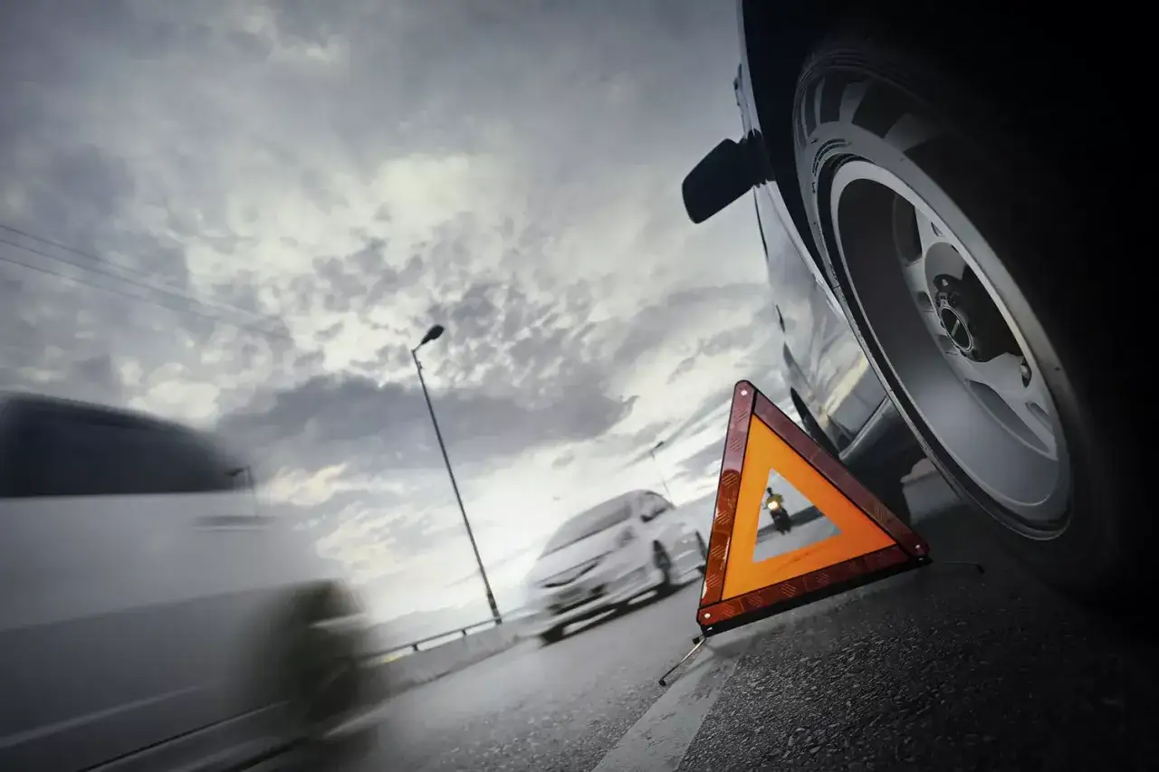 Trójkąt ostrzegawczy przy kole Toyoty. W tle inne samochody i zachmurzone niebo. Pomyśl o **toyota assistance gwarancyjny** w razie awarii.