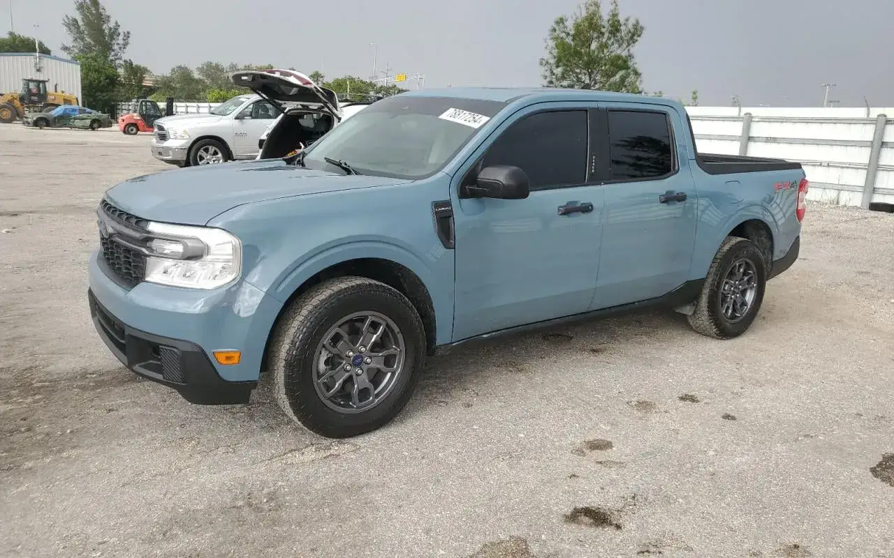 Niebieski Ford Maverick stoi na placu. W tle widać inne pojazdy i ogrodzenie.