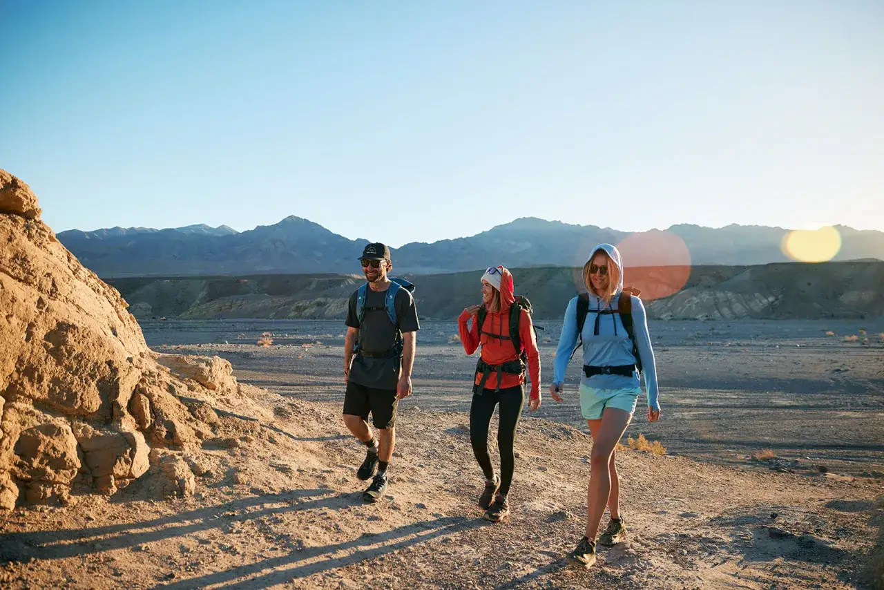 Trzech turystów wędruje przez pustynny krajobraz. Kobieta w środku ma na sobie czerwoną bluzę z kapturem, a jej towarzyszka po prawej jasnoniebieską. Wszyscy mają plecaki, a ich ubrania wykonane są z oddychającej wełny z merynosa.