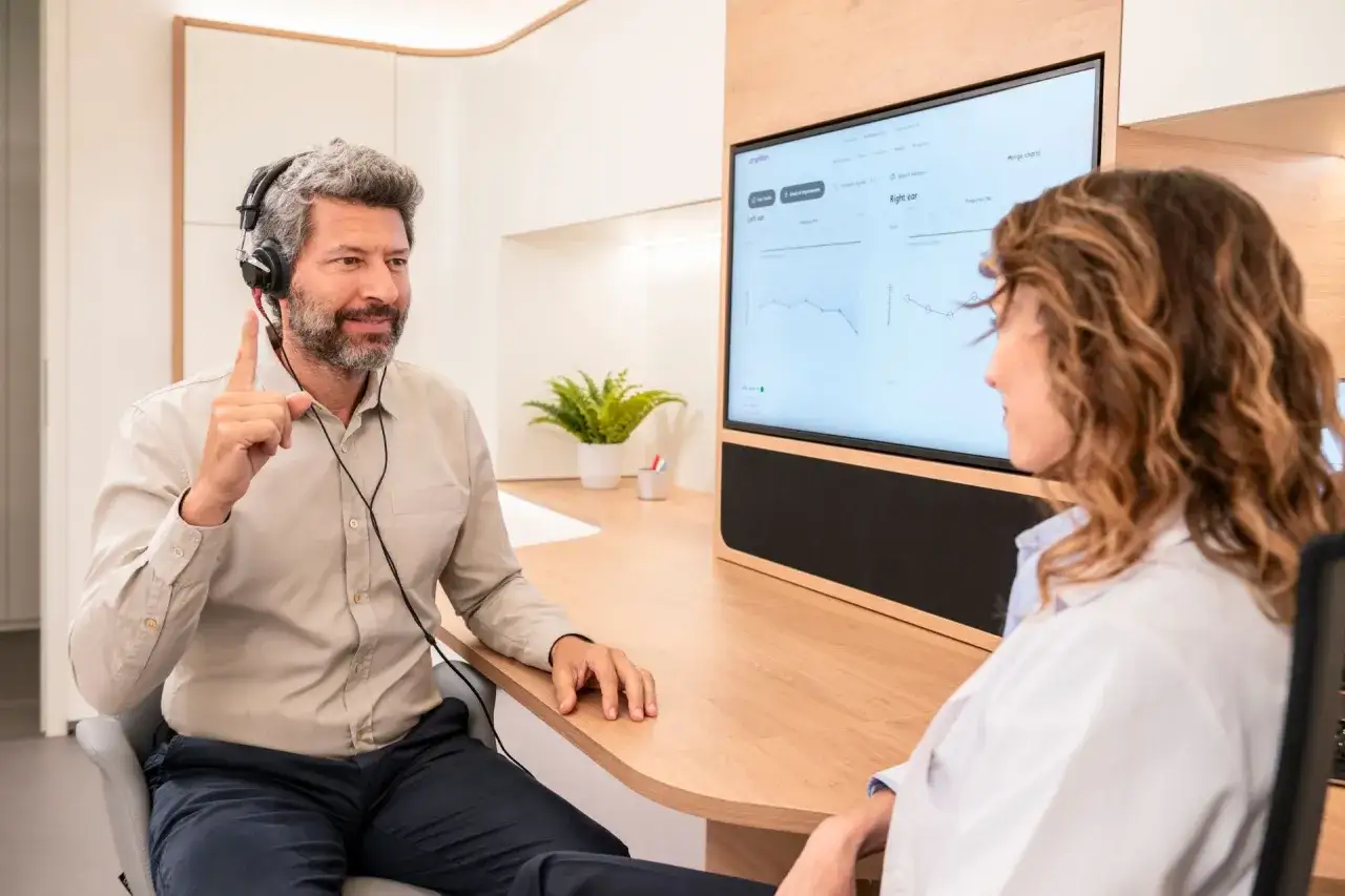 Mężczyzna w słuchawkach z podniesionym palcem rozmawia z audiologiem. Na ekranie wykresy audiometryczne. Dowiedz się, gdzie kupić aparat słuchowy.