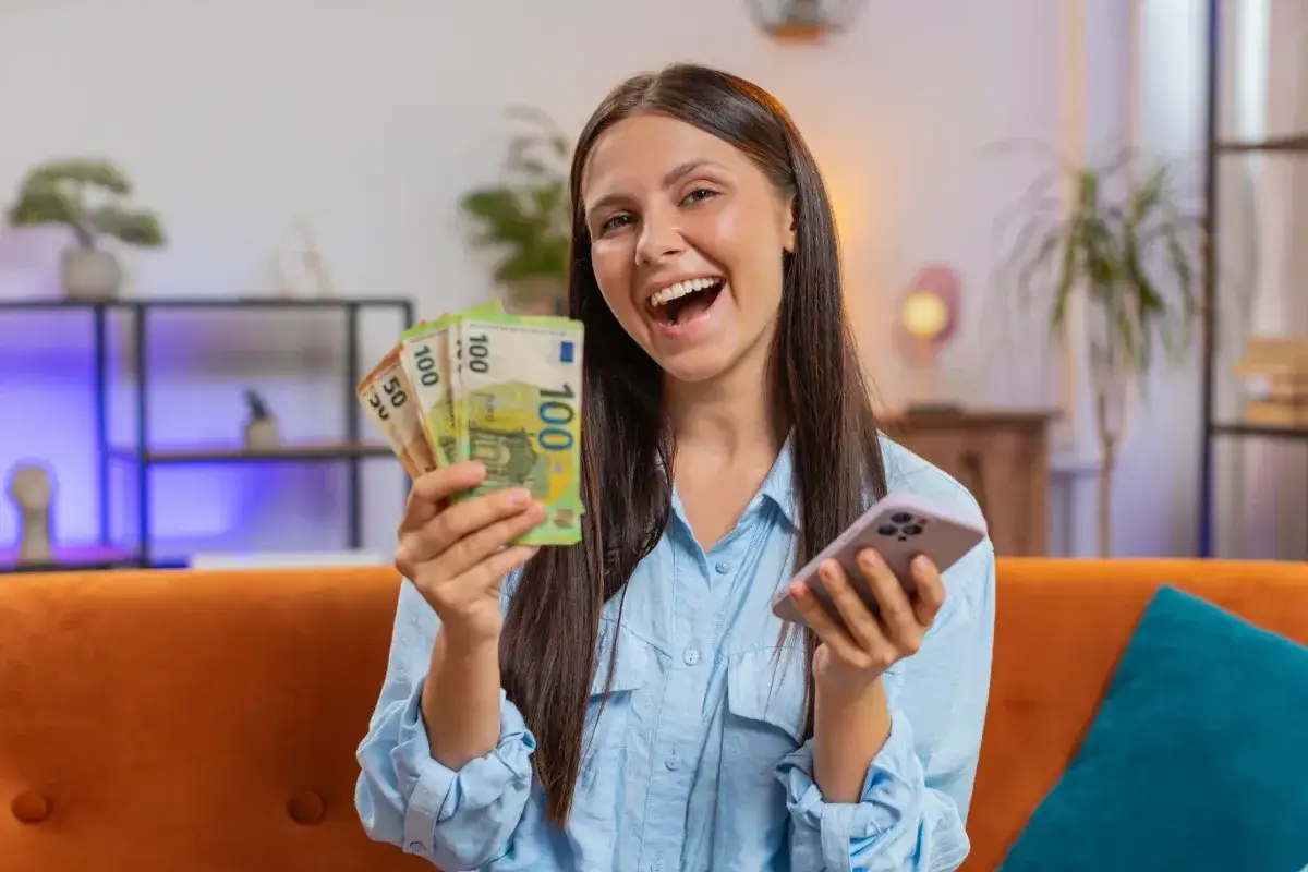 Uśmiechnięta kobieta trzyma banknoty euro i telefon, pokazując, na jakich aplikacjach można zarobić.