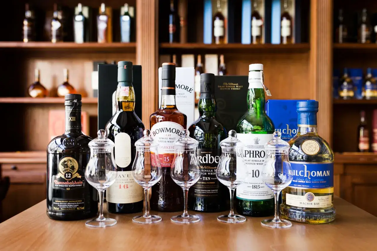 Rząd butelek whisky na drewnianym stole, w tym Bunnahabhain, Bowmore, Laphroaig i Kilchoman. Idealne na tanie dobre whisky.