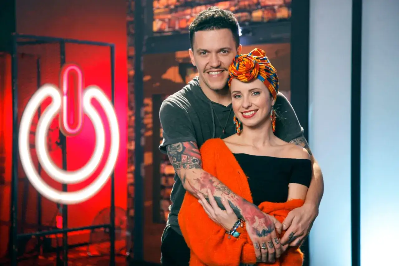 Uśmiechnięci uczestnicy programu, tworzący power couple, pozują na tle neonowego symbolu 