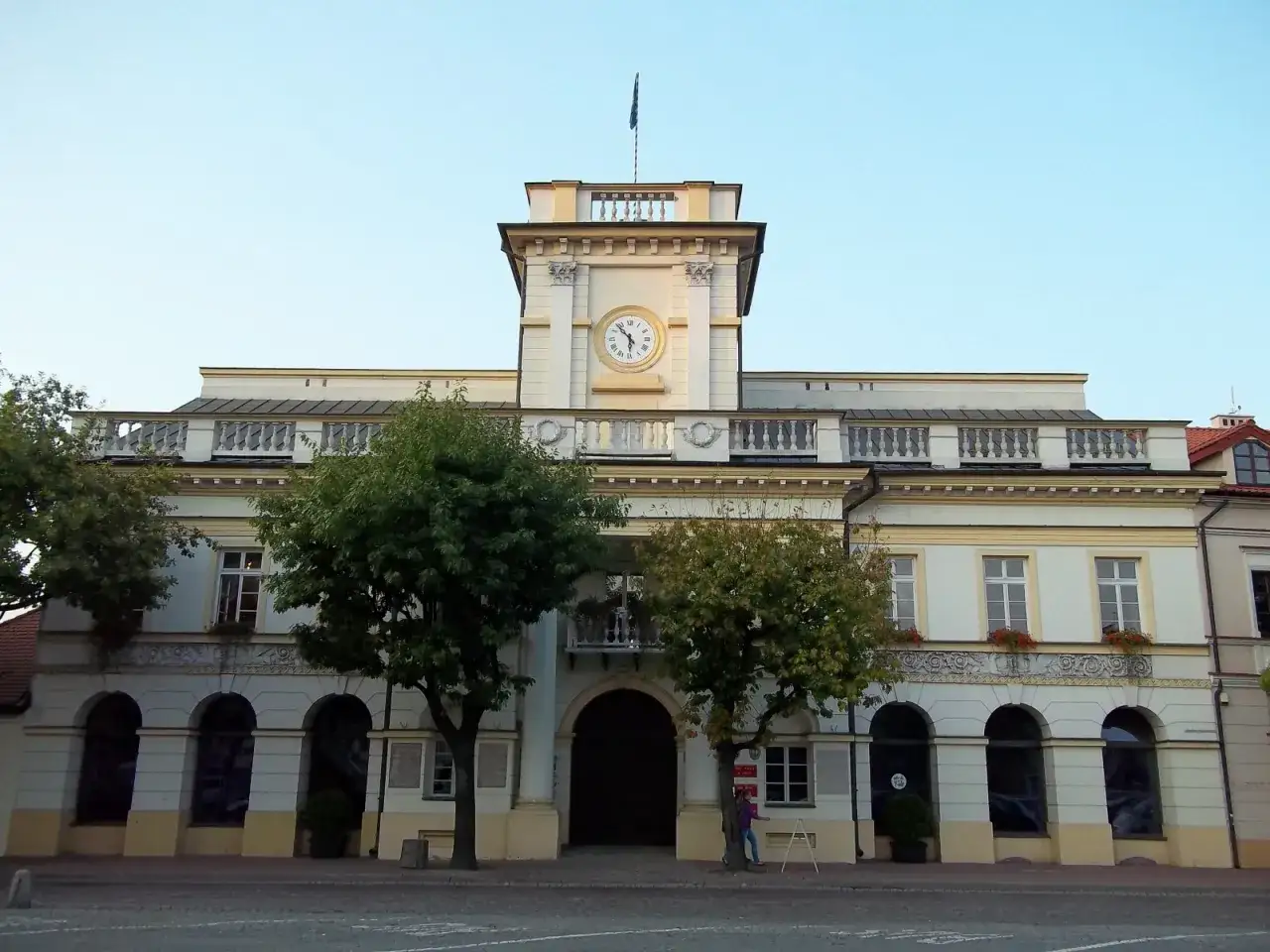 Stary rynek w Łowiczu. Ratusz z zegarem na wieży, otoczony drzewami, z łukowymi arkadami i balustradą.