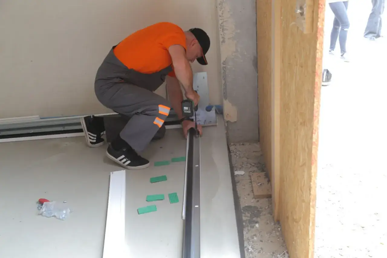 Majster w pomarańczowej bluzie i szarym ogrodniczkach przykręca prowadnicę, by uszczelnić drzwi do garażu.