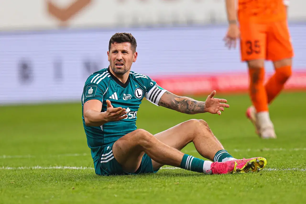 Piłkarz Legii Warszawa siedzi na murawie, gestykulując. W tle widać zawodnika Pogoni Szczecin.