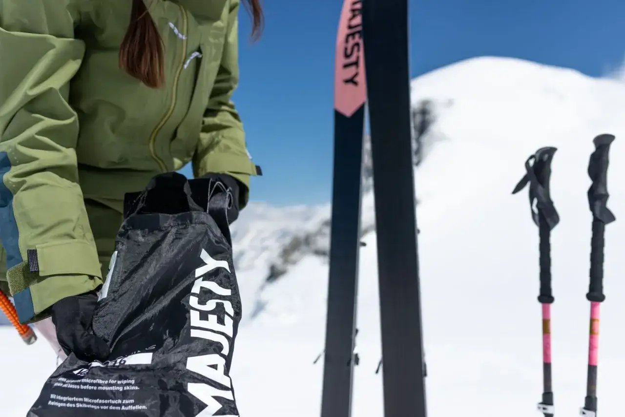 Osoba w zielonej kurtce trzyma czarną torbę z logo Majesty. Obok stoją narty i kije. Zastanawiasz się, ile kosztują narty?