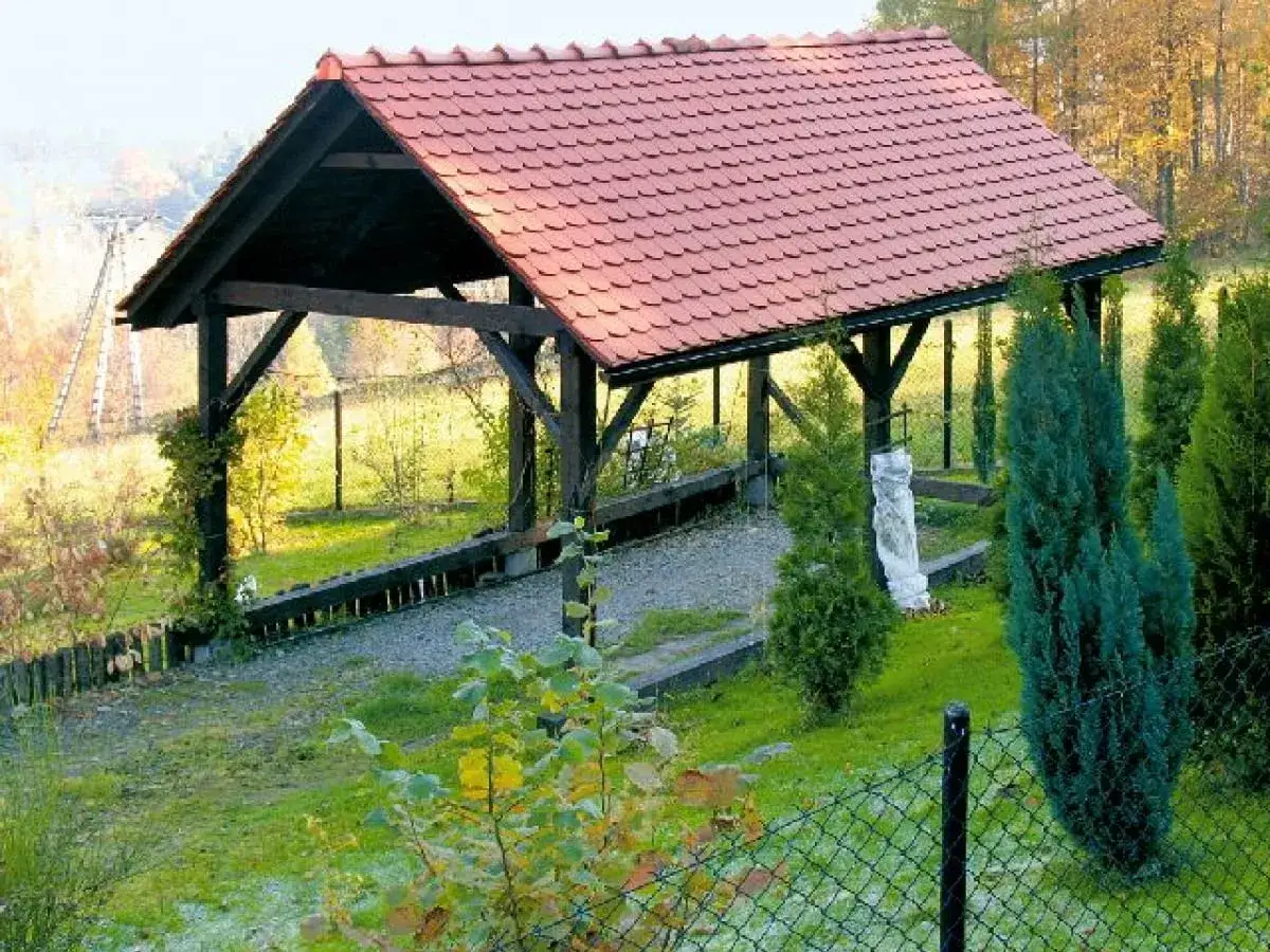 Drewniana wiata z czerwonym dachem, służąca jako garaż, stoi na tle jesiennego krajobrazu.