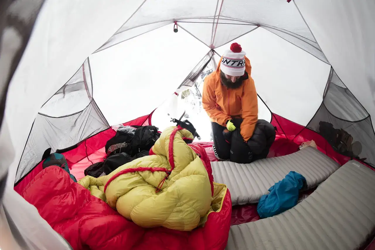 W namiocie, gdzie temperatura wynosi 10 stopni, mężczyzna w pomarańczowej kurtce przygotowuje się do spania. Wok&oacute;ł śpiwory i materace.