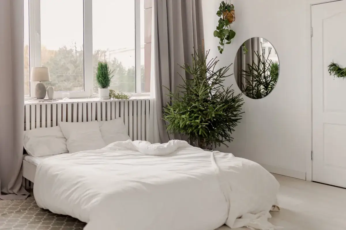 Sypialnia z białymi meblami i pościelą. Jasne, białe ściany tworzą przestronne wnętrze. W rogu stoi choinka.