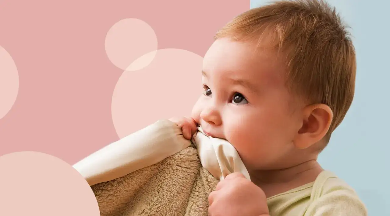 Maluch gryzie kocyk, być może z powodu ząbkowania. Jego oczy patrzą w g&oacute;rę, a tło jest w pastelowych kolorach.